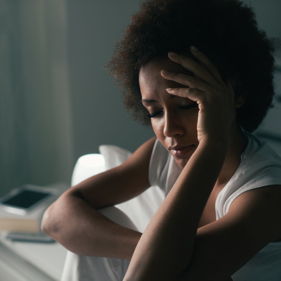 woman sits near window on bed hand holds hand to forehead with eyes closed 
